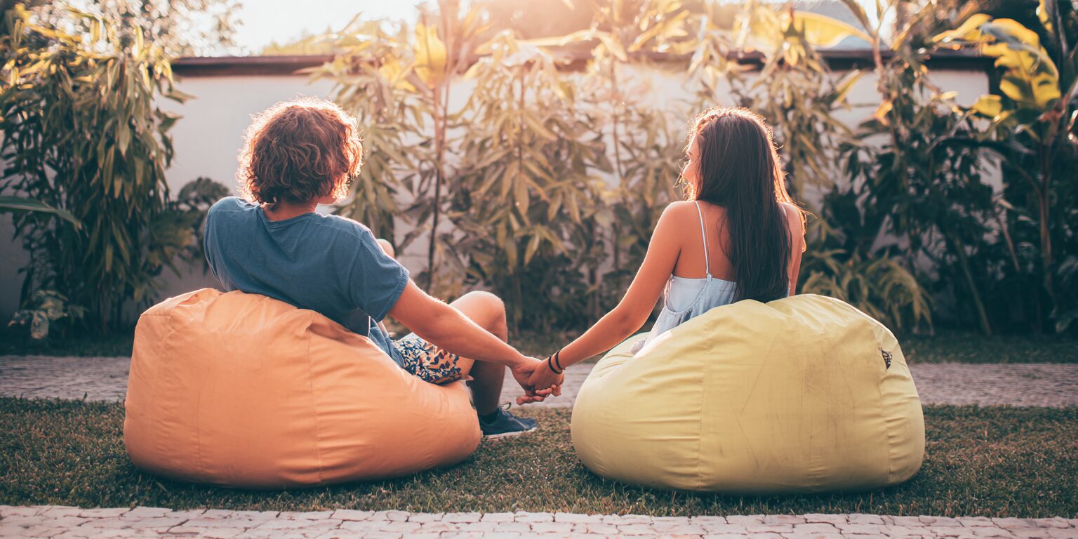 Mladý pár oddychuje na dvoch sedacích vakoch v záhrade, držia sa za ruky a užívajú si letnú pohodu.